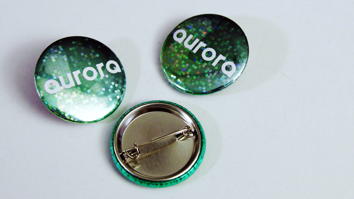 Green glittery background with white Auroa logo on a 32mm (1.25 inch) button badge