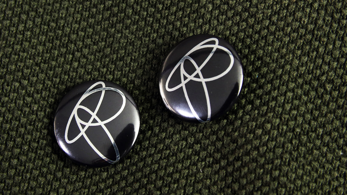 R-monogram logo printed black and metallic silver on a small 25mm (1-inch) sized button badge