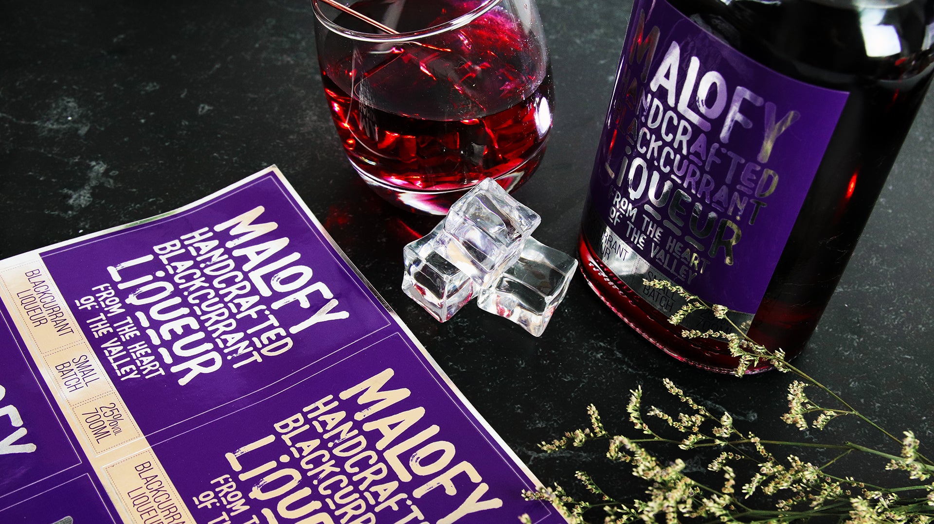 Rectangle mirror silver branding label applied to a glass bottle containing blackcurrant liqueur with a stack of sticker sheets next to a tilted glass
