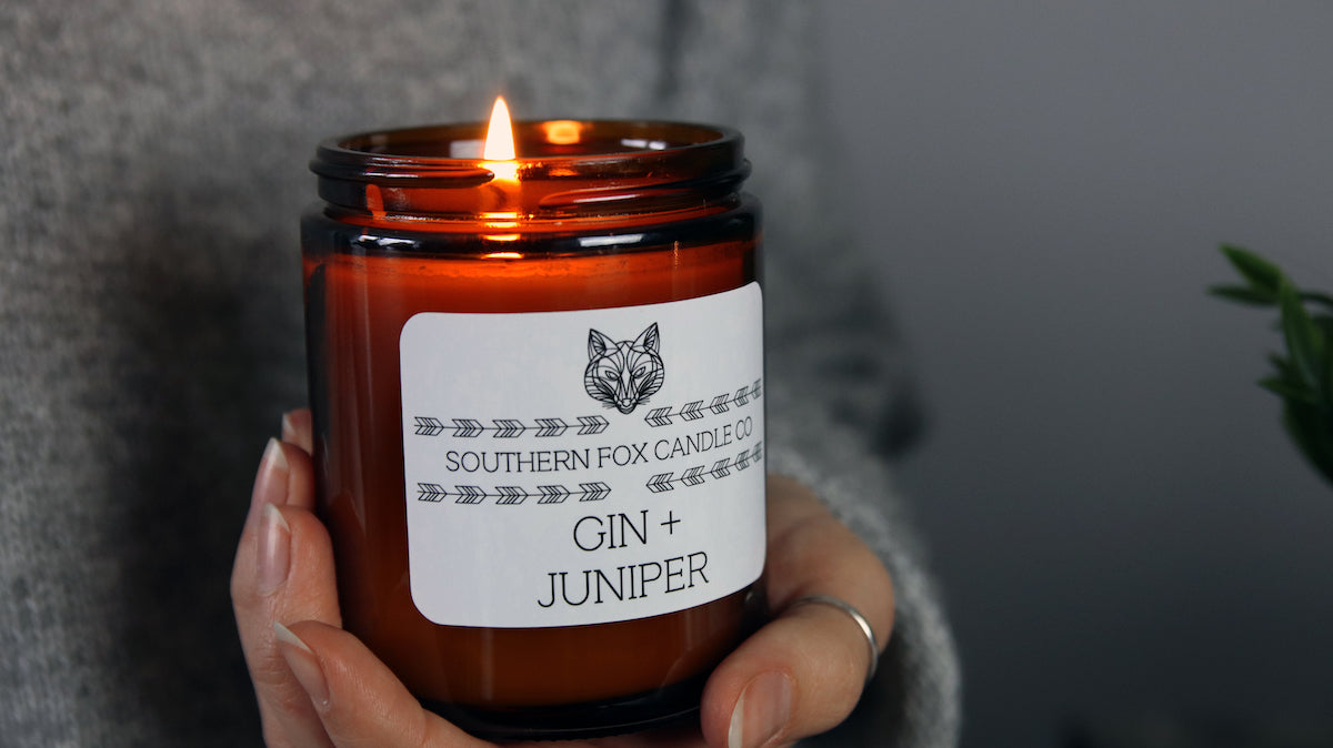 White vinyl label with rounded corners applied to an amber gin and juniper candle jar that is being held