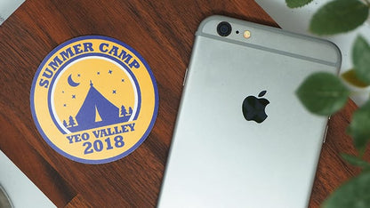 Circle biodegradable paper stickers with summer camp logo next to an iPhone