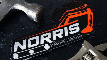 Die cut heavy duty sticker sample with norris plant hire and sales logo on a black table next to tools
