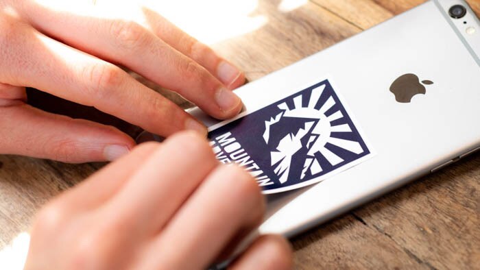 Rectangular white vinyl sticker with mountain adventure logo applied to a silver iPhone