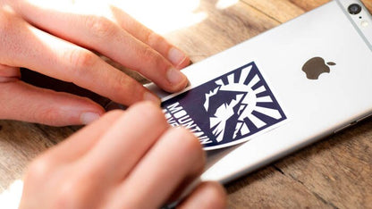 Rectangular white vinyl sticker with mountain adventure logo applied to a silver iPhone